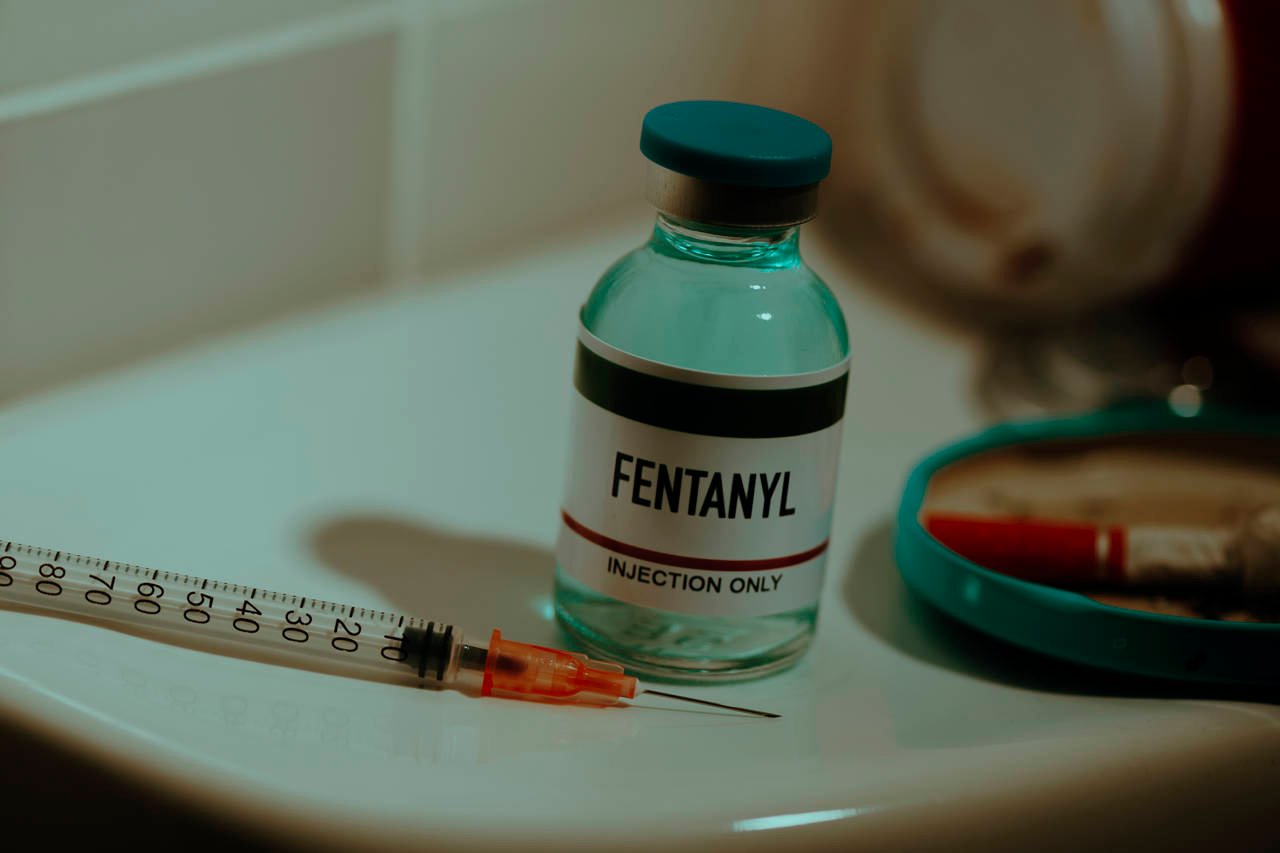 a simulated vial of fentanyl and a syringe on the cistern of a toilet in a restroom next to a cigarette butt